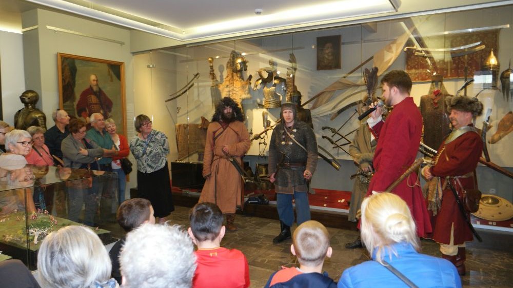 A few men in historical costumes talk to a group of people. Behind them are various exhibits behind a glass wall.