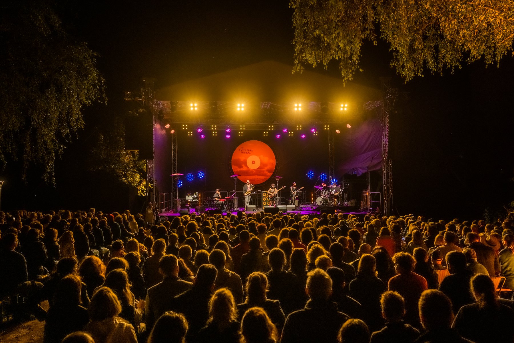 Crowd in front of an outdoor stage. It's already dark, people's heads are illuminated by orange light. - grafika artykułu