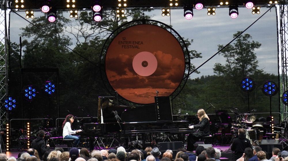 Woman and man playing pianos on stage. People in front of stage, treetops visible behind it.