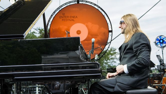 A smiling Leszek Możdżer is sitting at the piano. It's daytime, there's a big orange vinyl record at the back as stage decoration.