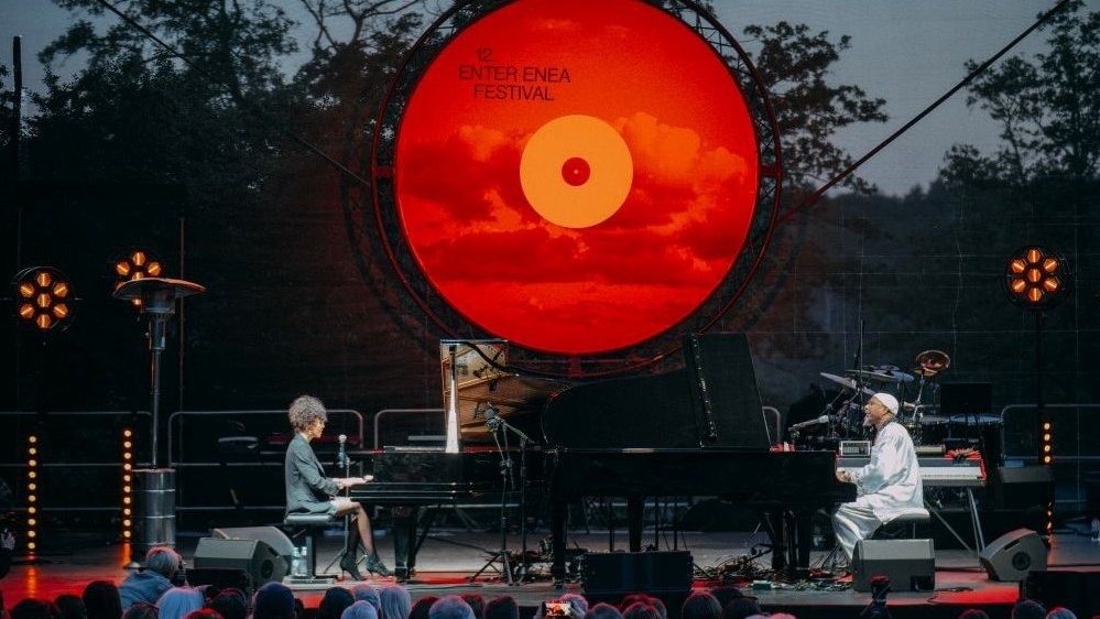 A woman and a man playing pianos on stage. It's getting dark, there's a big orange vinyl record at the back as stage decoration..