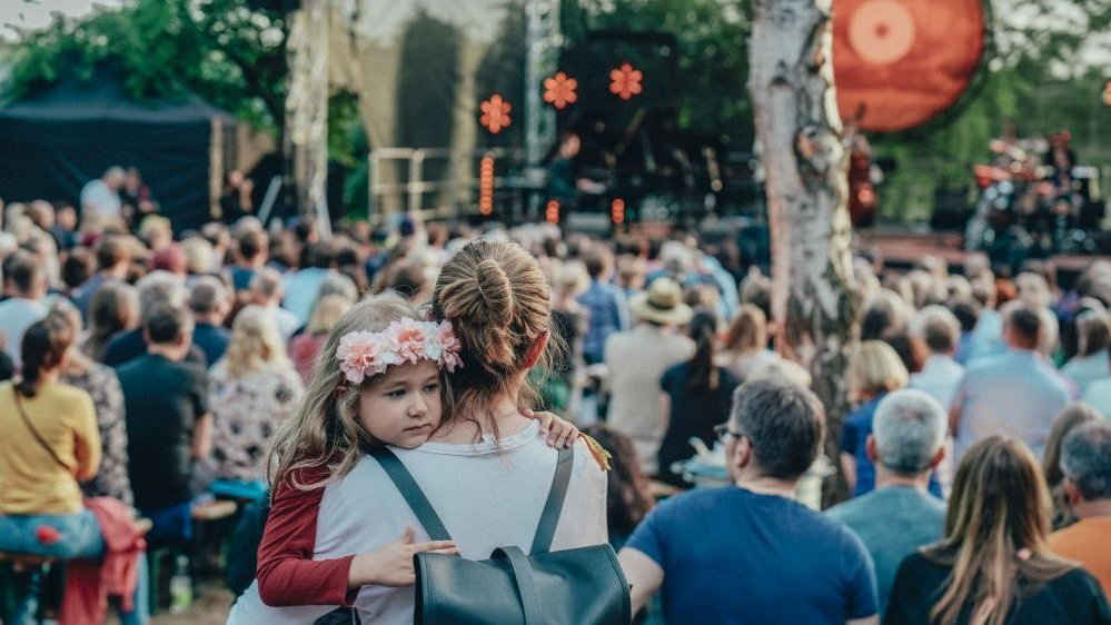 A colorful crowd of people in front of the outdoor stage. In the foreground, a mother holds her daughter with a flower wreath on her head.