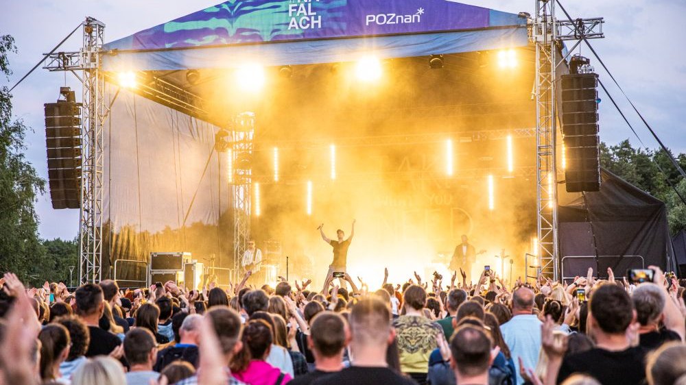 An outdoor stage lit with yellow light. A band is playing on stage, a colorful crowd is in front of it.