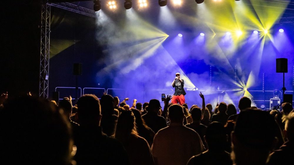 A man performing on stage, the stage lit with blue light. A crowd of people in front of the stage.