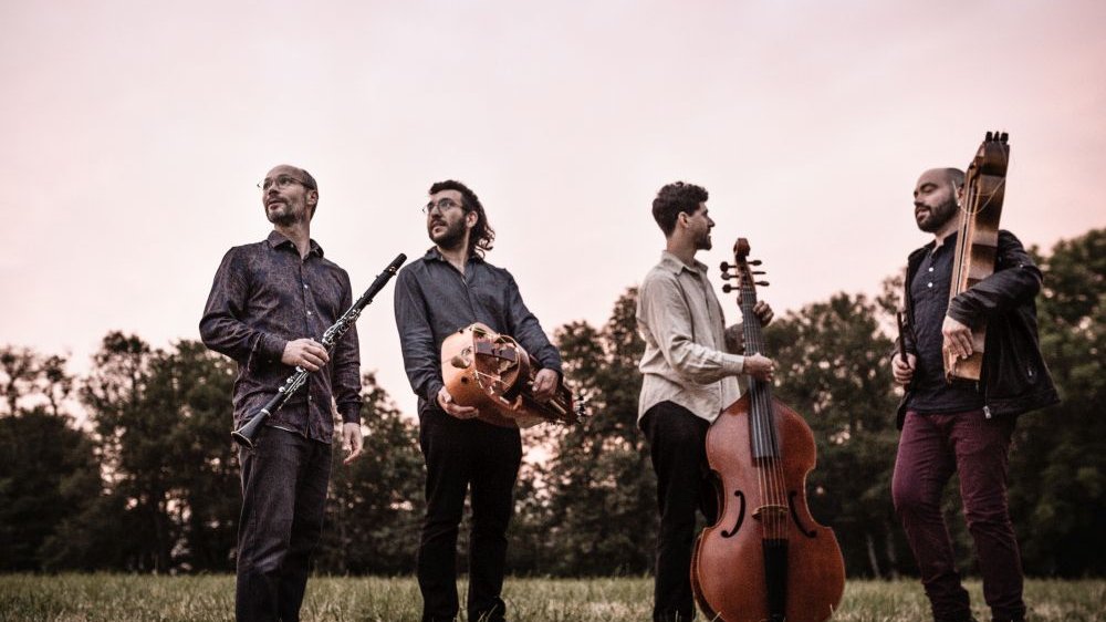 Four men hold instruments, including a cello and a clarinet. They are standing in the middle of a meadow.
