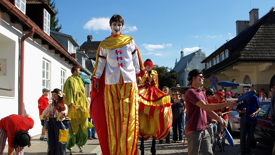 A colorful procession through the city. In the foreground, a colorfully dressed figure on stilts.