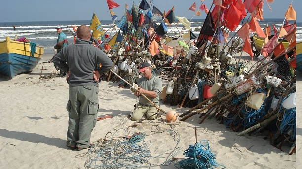 Seaside beach. On the shore, two men by a boat, colorful flags behind them.