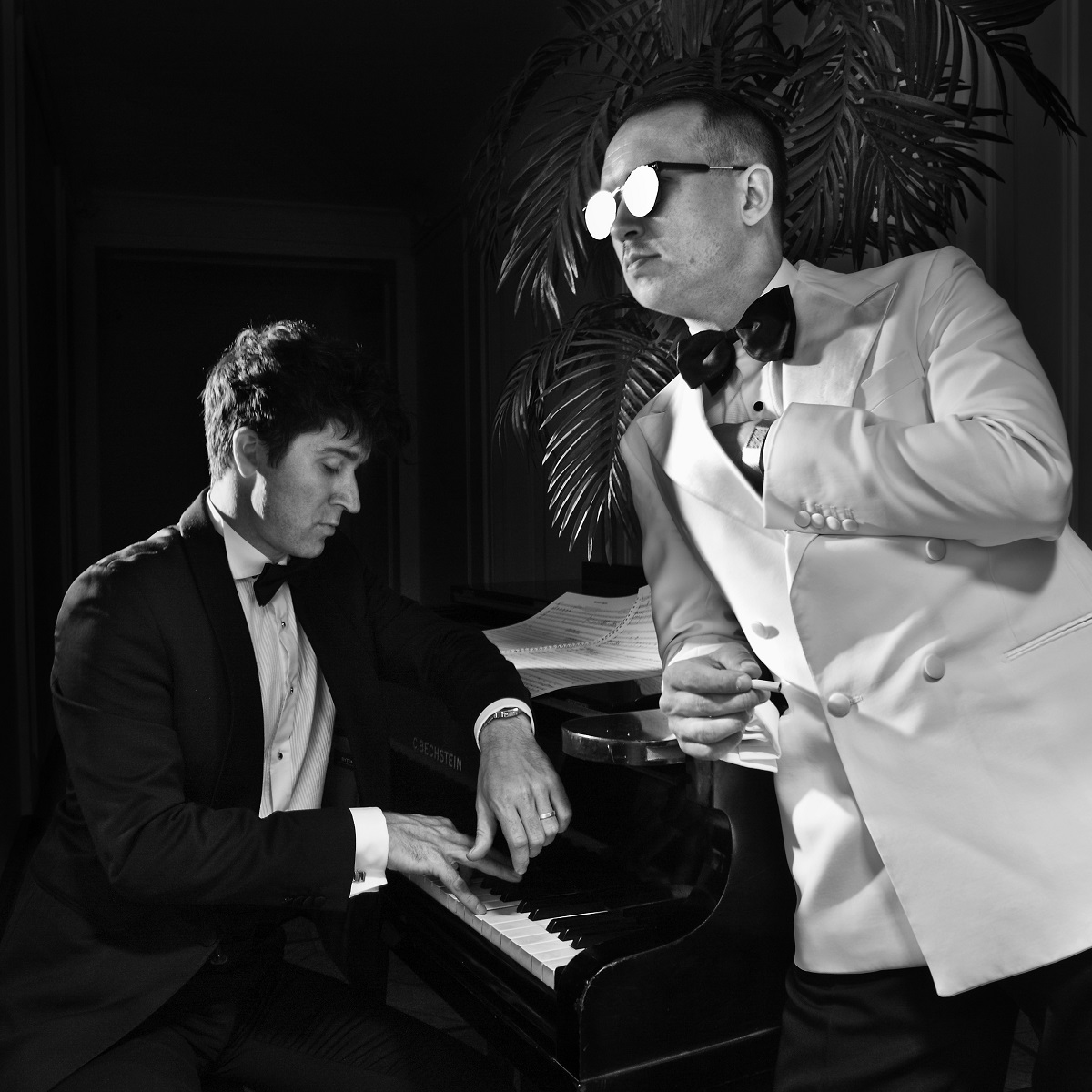 Black and white photo of two men: one sitting at the piano, another standing by it, leaning on it with his arm. - grafika artykułu