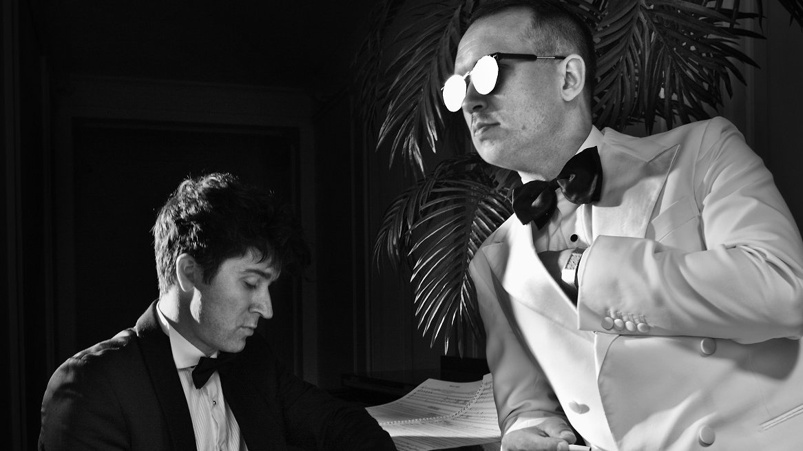 Black and white photo of two men: one sitting at the piano, another standing by it, leaning on it with his arm.
