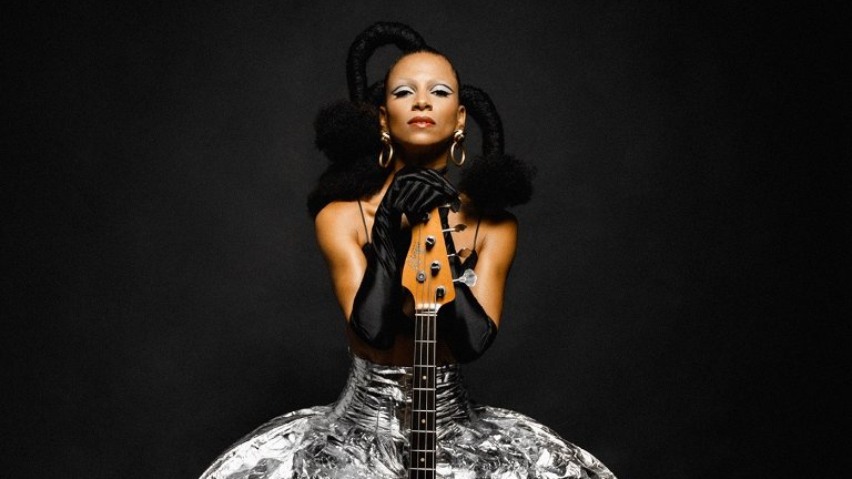Photo of a woman in silver dress, sitting behind a guitar. Black background.