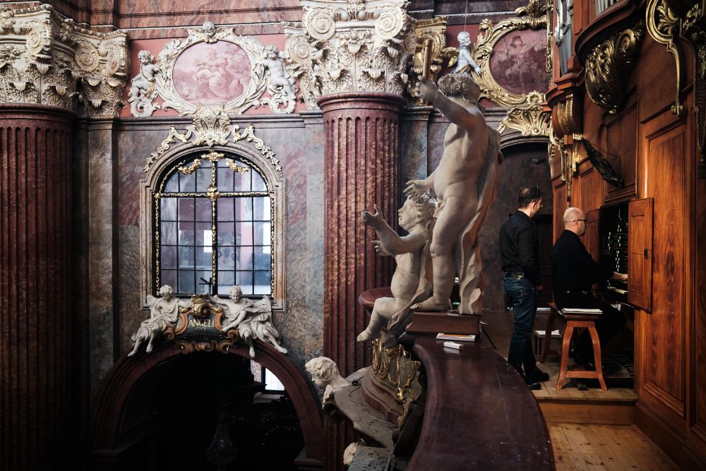 The interior of the parish church. At the top, by the side organs, sits a musician, behind him stands his companion. On the balustrades stand beautiful figures of cherubs, on the walls around hang ornate paintings of the "Stations of the Cross". - grafika artykułu