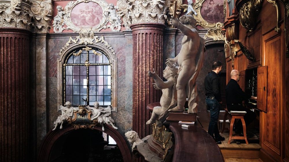 The interior of the parish church. At the top, by the side organs, sits a musician, behind him stands his companion. On the balustrades stand beautiful figures of cherubs, on the walls around hang ornate paintings of the "Stations of the Cross".
