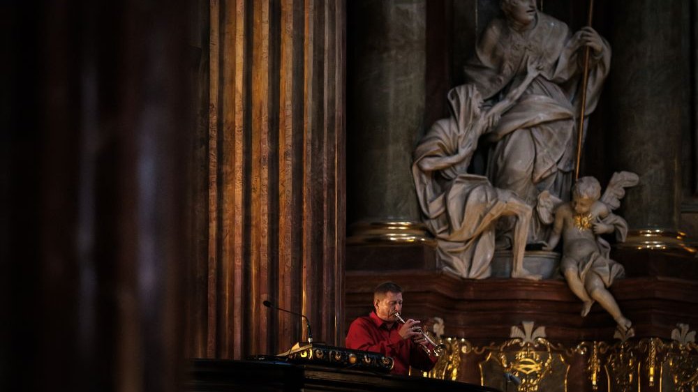 A musician in a red shirt plays a trumpet, standing to the side of the altar. Above him tower the sculptures in the parish church, depicting saints and cherubs.