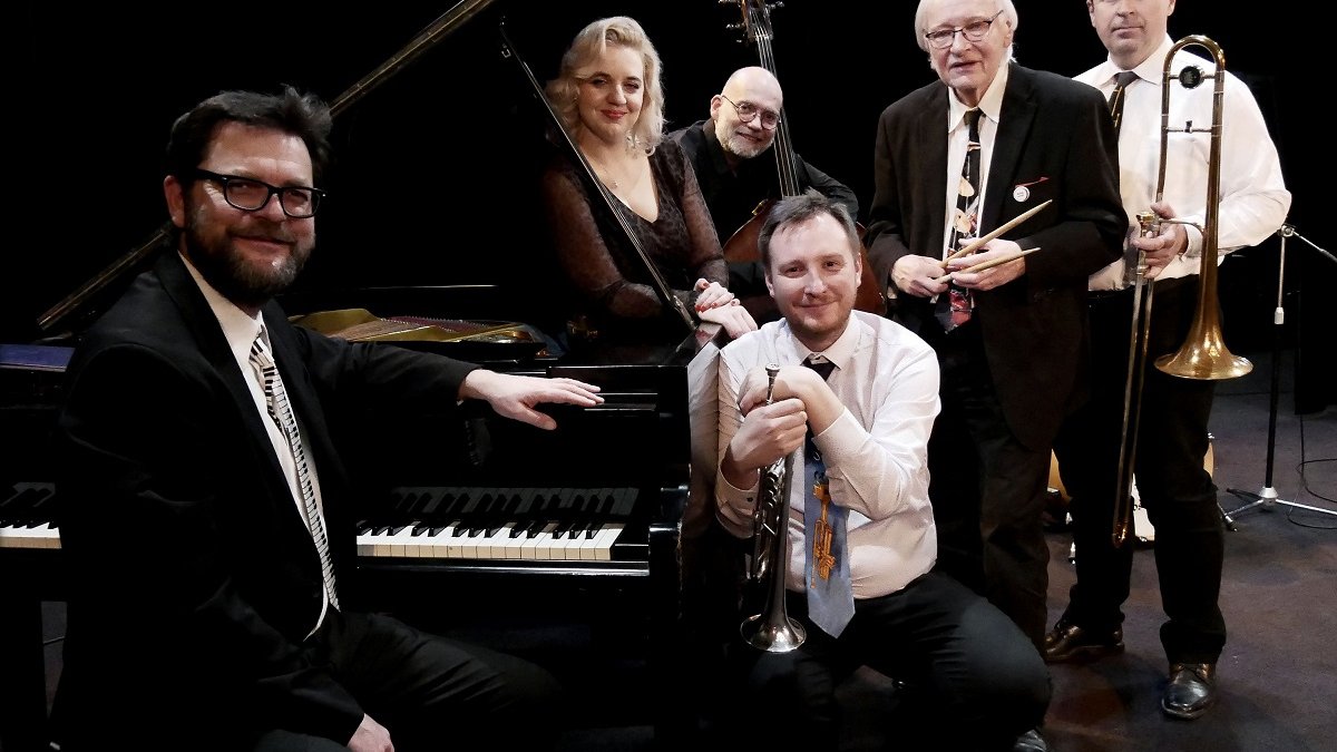 Photo of five men and one woman elegantly dressed. One of the men is sitting at a piano, the others are standing next to it. The men are holding musical instruments.