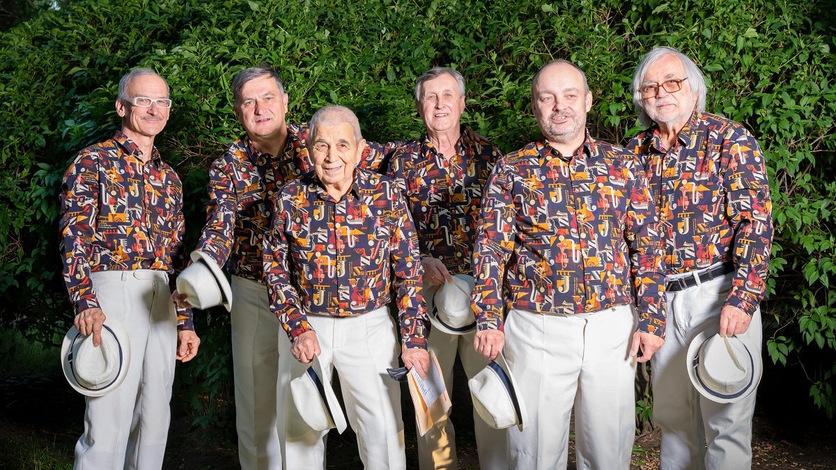 Photo of six older men wearing colorful shirts and white pants with white hats in their hands. Green bushes in the background.