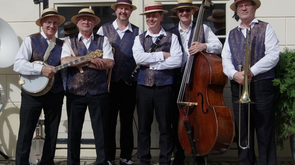 Six men dressed in white shirts, black trousers, blue vests and straw hats in front of a glass door. Each of them holds a musical instrument in his hands.