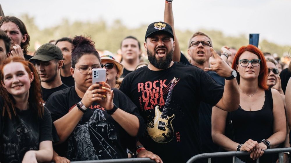 People in front of the stage. One of the men is wearing a festival T-shirt with a drawing of an electric guitar.