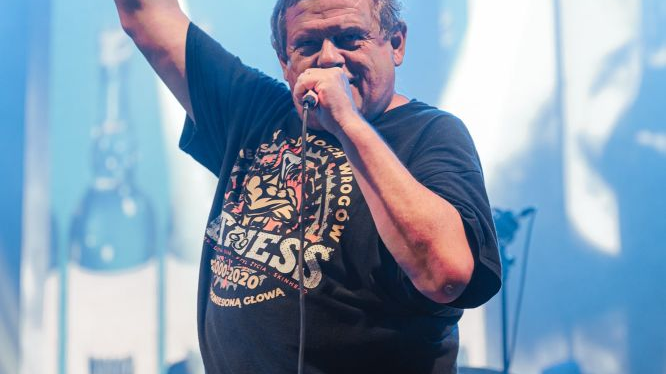 Kazik Staszewski during a performance on stage. He holds a microphone in one hand and raises the other up.