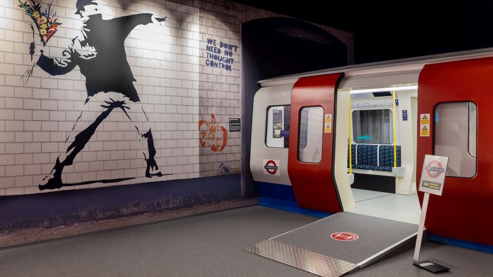 An exhibition installation with a red and white subway car and a drawing on the wall depicting a masked man throwing a bouquet of flowers.