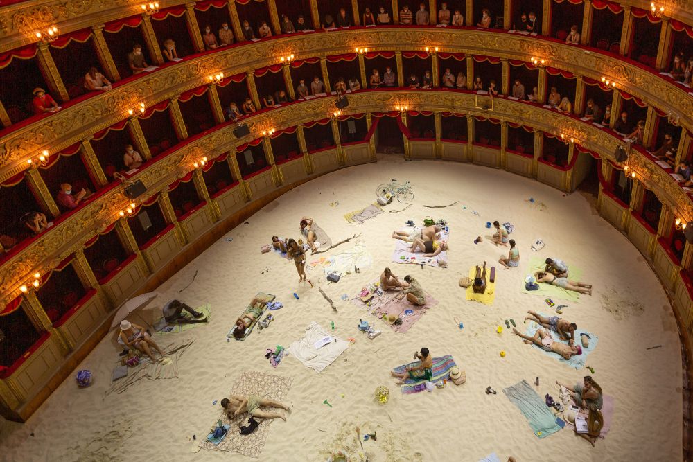 Classic opera interior, spectators on red and gold balconies. On the stage, a staged beach. There is a lot of sand, actors lie or sit on beach towels. - grafika artykułu