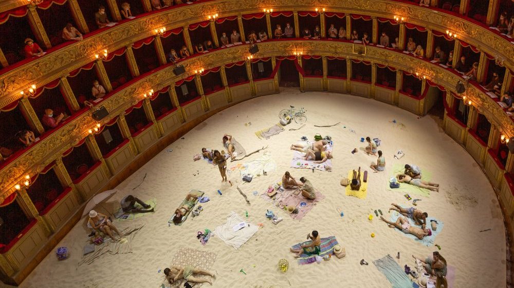 Classic opera interior, spectators on red and gold balconies. On the stage, a staged beach. There is a lot of sand, actors lie or sit on beach towels.