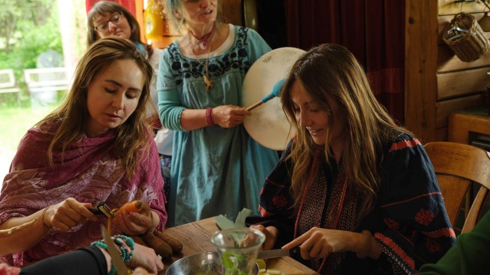 Six women in boho outfits in a wooden hut. One of them plays a drum, most of them peel vegetables at the table.