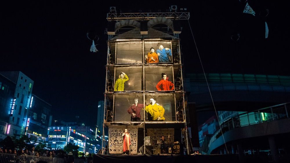 An outdoor set resembling a cross-section of a house with actors inside. It's night, and the city is dark all around.