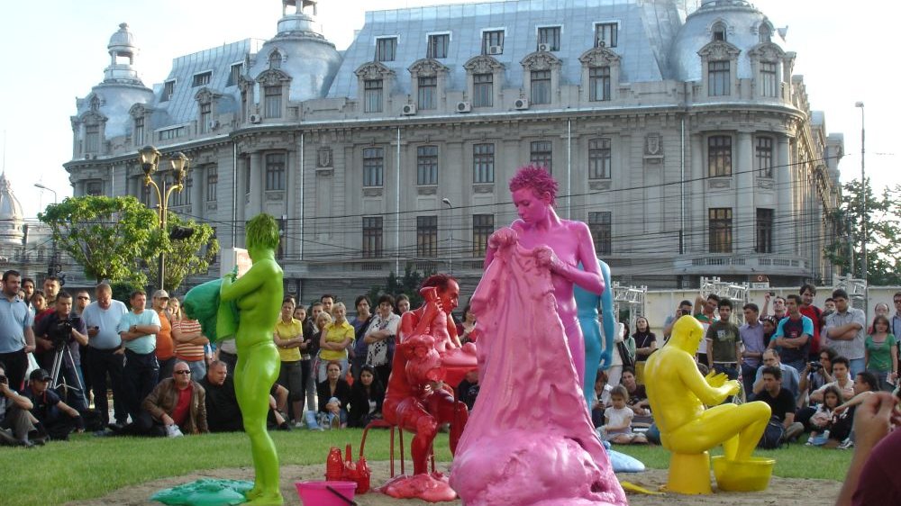 Painted green, red, yellow and pink, the actors perform an outdoor play among the people. Behind them is a stately, gray building.