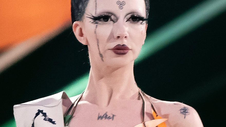 Portrait of a non-binary person with black stretchy pigtails. She is wearing a paper bra and a sash. She has a rainbow pin attached to her bra.