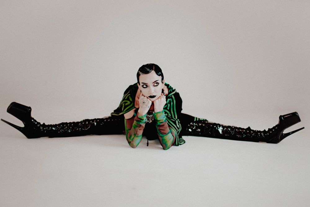 Photo of a dark-haired young person wearing long black boots, sitting on the floor with her legs spread wide, supporting her head with her hands. - grafika artykułu