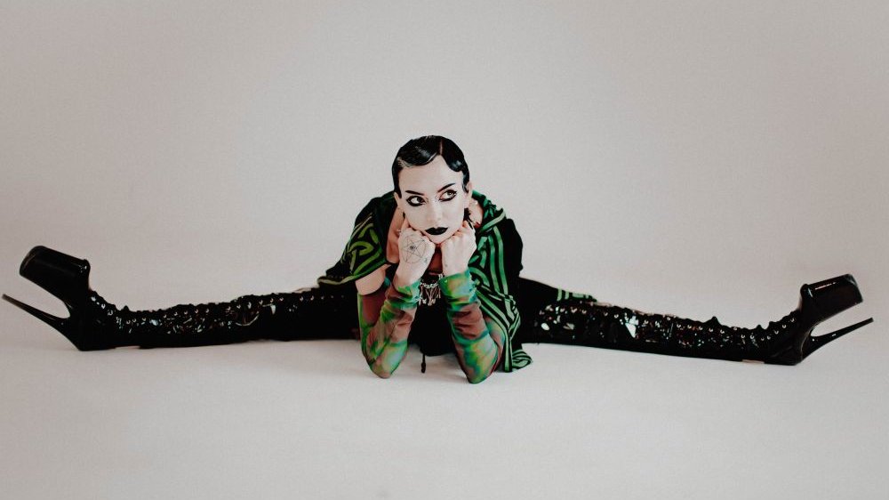 Photo of a dark-haired young person wearing long black boots, sitting on the floor with her legs spread wide, supporting her head with her hands.