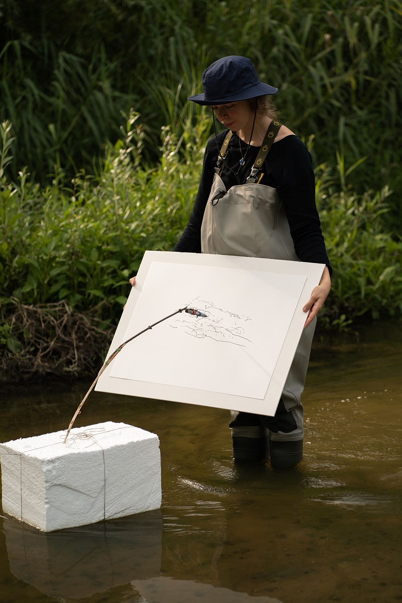 Photo of a woman, who stands in a river and holds a board with a sheet of paper. Oposite her, there is a construction consisting of a white box and a kind of metalic pointer touching the board with a paper. - grafika artykułu