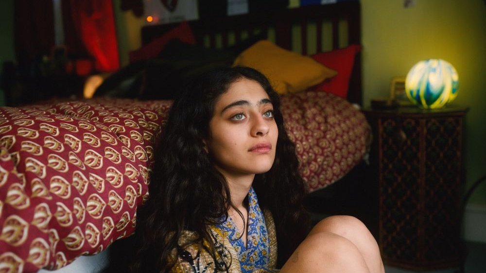 A young girl with big green eyes and dark curly hair sits on the floor in a room. Her back is against the bed.