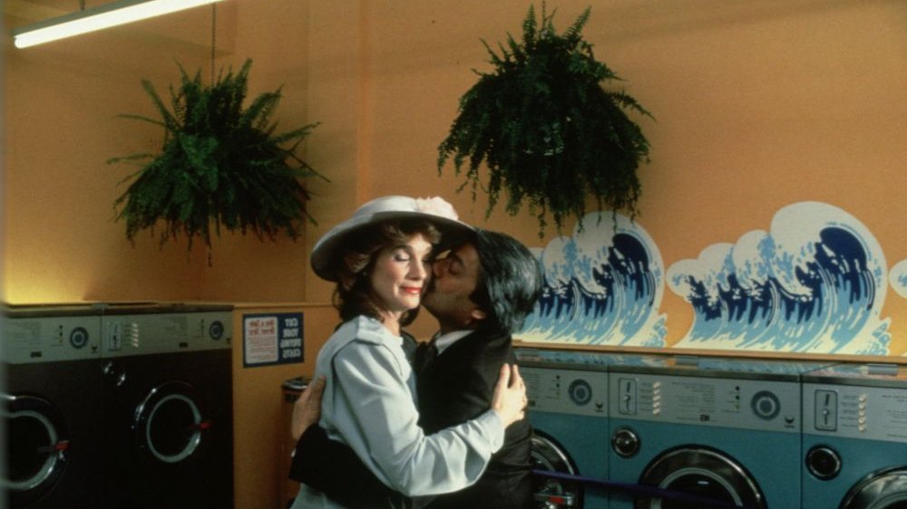 An elegantly dressed man and a woman in a hat hug. Behind them, rows of washing machines. A picture of choppy waves is painted on the wall.