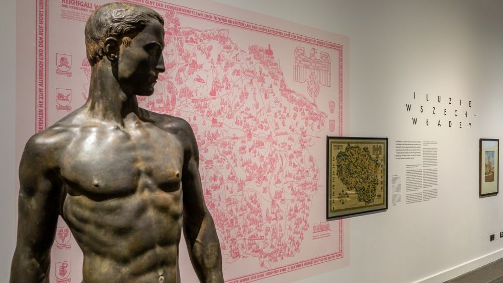 In the foreground there is a sculpture of a naked man, in the background on the wall there is, among other things, a pink city map.