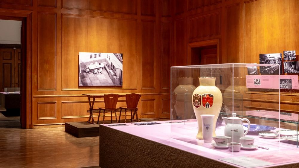 Exhibition hall. In the foreground, a glass display case with porcelain dishes inside. In the background, black and white photos on the walls.