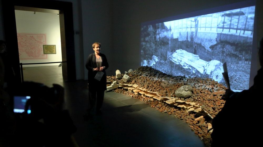 An exhibition hall in semi-darkness. A photograph is displayed on the wall, with stones arranged underneath. A woman stands in the middle of the room.