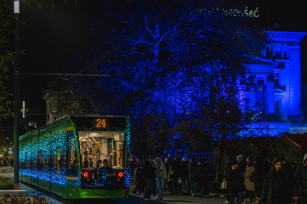 A tram decorated with Christmas lights, with people walking next to it. In the background, a tree and a building, illuminated by blue light. - grafika artykułu