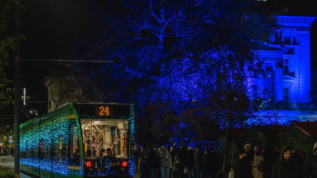 A tram decorated with Christmas lights, with people walking next to it. In the background, a tree and a building, illuminated by blue light.