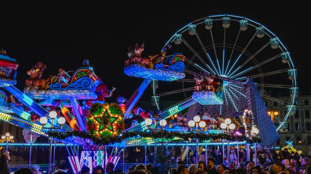 A colorful carousel, illuminated by many lights, on Plac Wolności. Each carriage has two funny reindeer. Behind the carousel, you can see a huge illuminated wheel - a Ferris wheel.