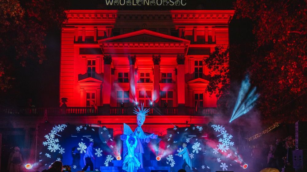 The Arkadia building illuminated in red from the side of Plac Wolności. A performance is taking place on the stage, with blue figures and decorations covered with large cut-out snowflakes.