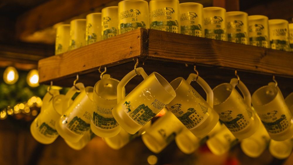 A wooden shelf on which many mugs and tankards stand or hang with the inscription "Betlejem Poznańskie" and painted historic tenement houses from the Old Market Square.