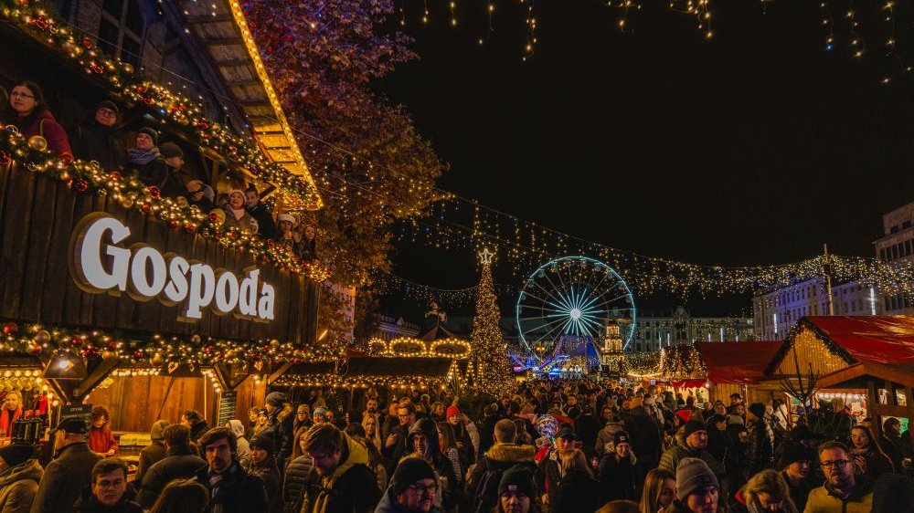 A crowd of people at the Bethlehem Poznańskie fair, in the distance you can see the Ferris wheel, on the left the Inn. All around stands are lit with lamps. It is evening or early night.