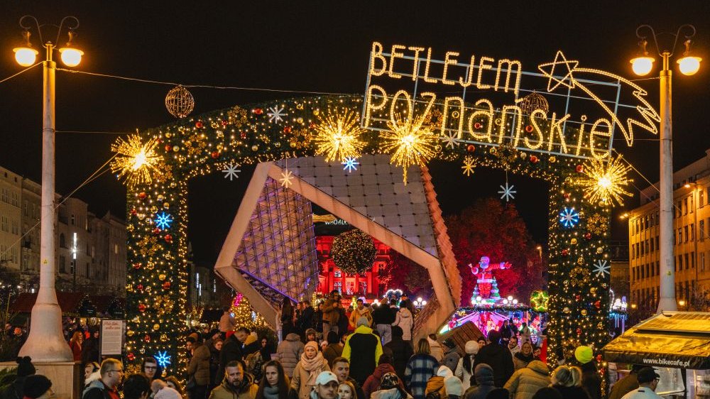 The entrance to the fair, decorated with lights, Christmas balls and the inscription "Betlejem Poznańskie", and people walking around it. Behind the entrance is a modern geometric structure, which is normally a fountain.