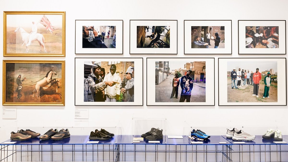 A display of sneakers - several pairs on a long rack against the wall. Above the stand with sneakers, on the wall, photos mostly of dark-skinned men.