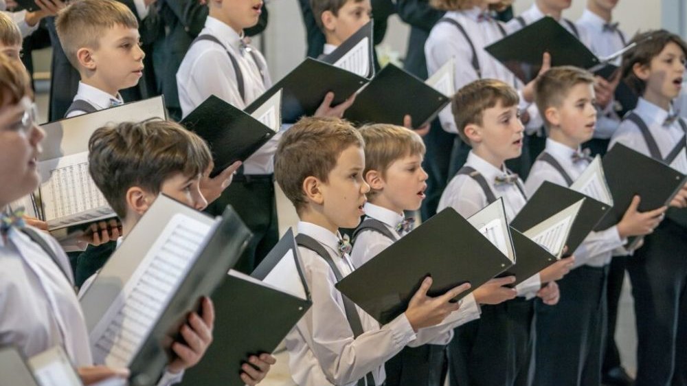 A group of singing boys. The boys are dressed identically in dark trousers, white shirts and suspenders. Each of them holds a songbook in their hands