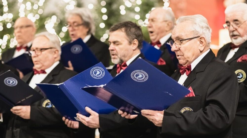 Photo of several men singing. The men are dressed in suits with red bow ties, holding songbooks in their hands.