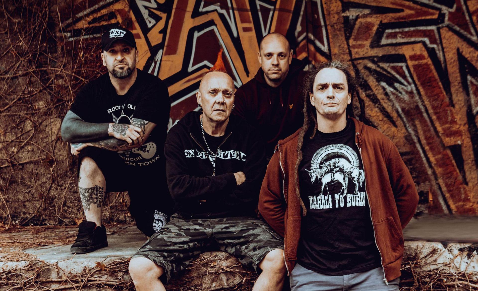 Photo of The Exploited band - four men in dark clothes; wall with graffiti as a background. - grafika artykułu