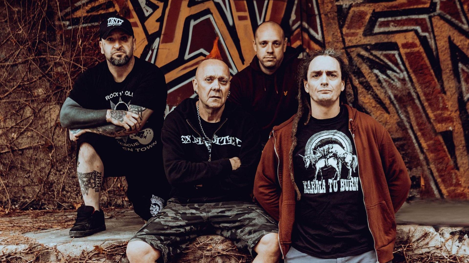 Photo of The Exploited band - four men in dark clothes; wall with graffiti as a background.