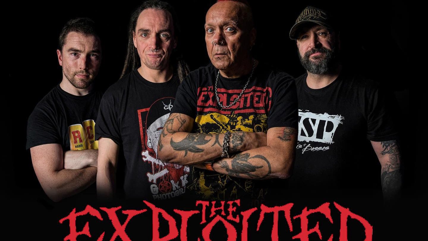 Photo of The Exploited band - four men in dark clothes; black background. At the bottom of the photo the name of the band.